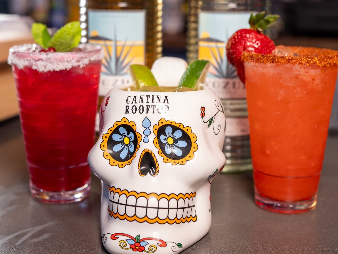 Margaritas and a Cantina skull mug in front of Vozul tequila bottles